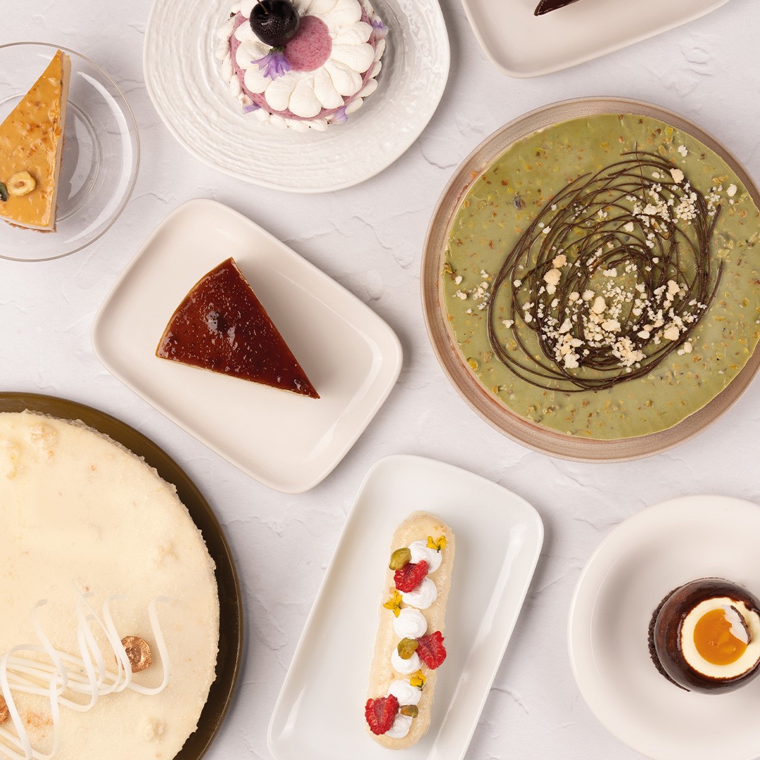 Overhead view of assorted desserts including cheesecake, flan, pistachio tart, eclair, and chocolate mousse cups on w...
